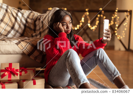 Unhappy young black woman looking at smartphone screen, sitting on floor near Christmas gift boxes at home 83406067