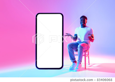 Positive black man sitting on chair near huge cellphone with empty white screen in neon light, mockup for mobile app 83406068