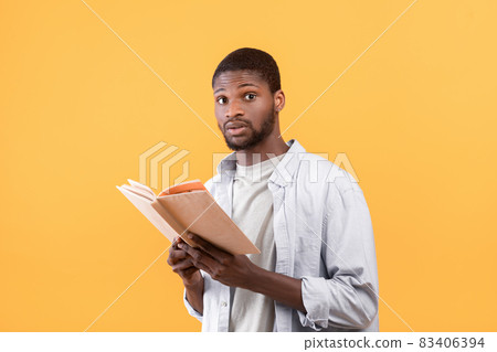 Shocked black male student holding textbook, looking at camera with shock over yellow studio background Shocked black male student holding textbook, looking at camera with shock over yellow studio background 83406394