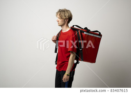 Portrait of a young courier, delivery man in red uniform with a thermal backpack isolated on a white background. Fast home delivery. Online order. Courier delivers groceries home. 83407615
