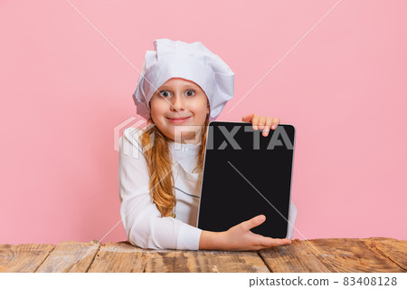 Portrait of little cute girl in white cook uniform and huge chef's hat looking at camera isolated on pink studio background. 83408128