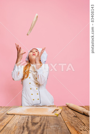 Portrait of little beautiful girl in white cook uniform and huge chef's hat at kids kitchen isolated on pink studio background. 83408143