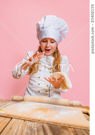 Portrait of little beautiful girl in white cook uniform and huge chef's hat at kids kitchen isolated on pink studio background. 83408159