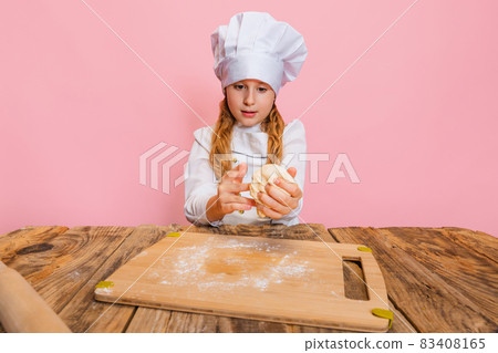 Portrait of little beautiful girl in white cook uniform and huge chef's hat at kids kitchen isolated on pink studio background. Portrait of little beautiful girl in white cook uniform and huge chef's hat at kids kitchen isolated on pink studio background. 83408165