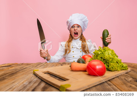 Young little girl in white cook uniform and huge chef's hat cutting fresh vegetables isolated on pink studio background. 83408169
