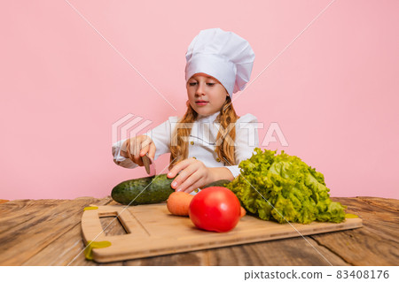 Young little girl in white cook uniform and huge chef's hat cutting fresh vegetables isolated on pink studio background. 83408176
