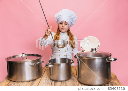 Young little girl in white cook uniform and huge chef's hat at kids kitchen with big pans isolated on pink studio background. Young little girl in white cook uniform and huge chef's hat at kids kitchen with big pans isolated on pink studio background. 83408210