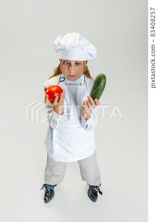 High angle view of little cute girl in white cook uniform and huge chef's hat posing isolated on white studio background. 83408257