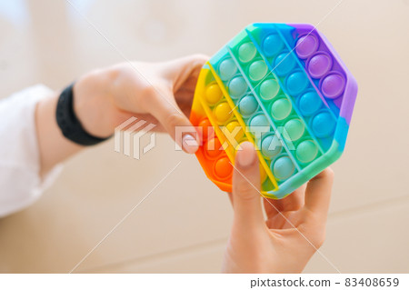 Close-up top view of of unrecognizable young woman playing with rainbow pop-it fidget toy sitting at table. 83408659