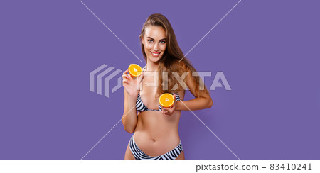 Slim cute girl in bikini holds halves of an orange while isolated on purple background. Summer time. Slim cute girl in bikini holds halves of an orange while isolated on purple background. Summer time. 83410241