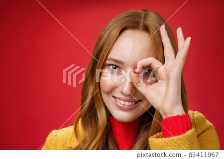 Zero worries only happiness. Portrait of charming cute and carefree redhead female with freckles and blue eyes smiling delighted showing okay gesture on eye and peeking at camera over red background Zero worries only happiness. Portrait of charming cute and carefree redhead female with freckles and blue eyes smiling delighted showing okay gesture on eye and peeking at camera over red background 83411967