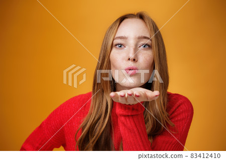 Close-up shot of sensual and attractive redhead female in red knitted outfit folding lips tilting forward as sending air kiss at camera giving mwah holding palm near mouth over orange wall 83412410