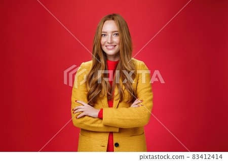 Charming successful young female in yellow autumn coat smiling broadly holding hands crossed on body in confident self-assured pose standing delighted and happy over red background 83412414
