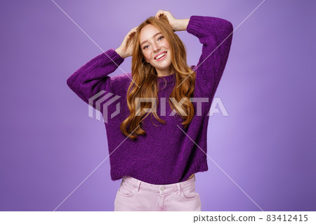 Carefree joyful and cool stylish redhead female student in purple warm sweater enjoying holidays stretching, holding hands near head, tilting and smiling happily relaxing against violet background 83412415