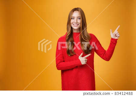 Friendly-looking joyfuly and energized redhead female in red sweater pointing at upper right corner promoting advertisement with broad delighted and tender smile posing against orange background 83412488