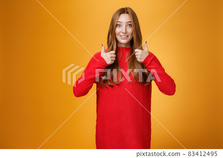 Excited and supportive redhead girlfriend asking opinion about new warm dress showing thumbs up and raising eyebrows questioned as waiting for opinion over orange background 83412495