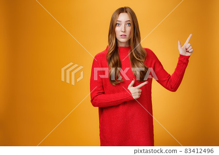 Portrait of elegant and stylish redhead 20s woman with freckles and long hair in knitted red dress pointing at upper right corner looking questioned and impressed, speechless over orange background 83412496