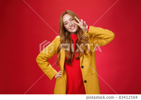 Friendly girl smiling happily, tilting head and showing peace or victory gesture near face, holding hand on hip as posing in stylish elegant yellow coat and sweater against red background Friendly girl smiling happily, tilting head and showing peace or victory gesture near face, holding hand on hip as posing in stylish elegant yellow coat and sweater against red background 83412504