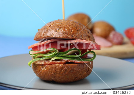 burger sandwich of buckwheat dark bun sausage salami vegetables cucumbers tomato on a gray plate on a blue background. Homemade instant food burger sandwich of buckwheat dark bun sausage salami vegetables cucumbers tomato on a gray plate on a blue background. Homemade instant food 83416283