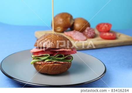 burger sandwich of buckwheat dark bun sausage salami vegetables cucumbers tomato on a gray plate on a blue background. Homemade instant food burger sandwich of buckwheat dark bun sausage salami vegetables cucumbers tomato on a gray plate on a blue background. Homemade instant food 83416284