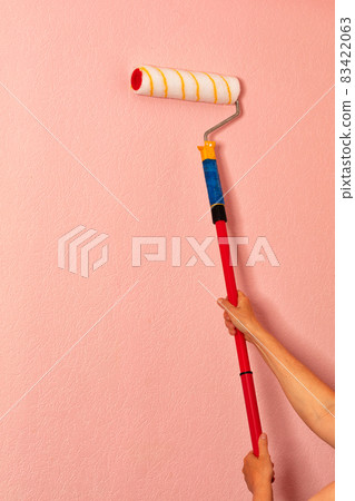 A fleecy paint roller in the female hands of a painter against the background of a pink wall. A fleecy paint roller in the female hands of a painter against the background of a pink wall. 83422063
