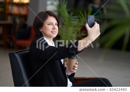 Portrait of young caucasian business woman wearing black suit doing selfie on the phone while coffee break Portrait of young caucasian business woman wearing black suit doing selfie on the phone while coffee break 83422493
