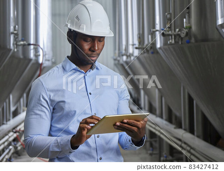 Portrait of a professional industrial engineer. Technician engineer in a safety helmet with a tablet. Large industrial plant background Portrait of a professional industrial engineer. Technician engineer in a safety helmet with a tablet. Large industrial plant background 83427412