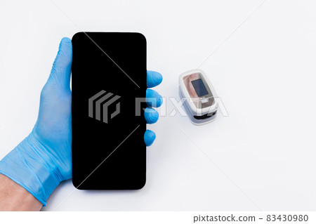 The hand of a medical doctor in a latex, rubber glove holds a mobile phone or smartphone with a screen against the background of the pulse oximeter The hand of a medical doctor in a latex, rubber glove holds a mobile phone or smartphone with a screen against the background of the pulse oximeter 83430980