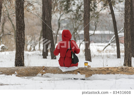 Girl in bright red winter jacket sits on log in winter snow-covered forest. Back view. Girl in bright red winter jacket sits on log in winter snow-covered forest. Back view. 83431608