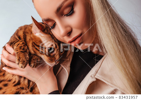 Portrait of an attractive cute young blonde woman with a bengal cat. Beautiful girl, looks at the cat, elegant hairstyle, impudent makeup, White background. Portrait of an attractive cute young blonde woman with a bengal cat. Beautiful girl, looks at the cat, elegant hairstyle, impudent makeup, White background. 83431957