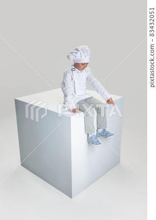 Studio shot of little school boy in white cook uniform and huge chef's hat sitting on huge box isolated on white studio background. 83432051