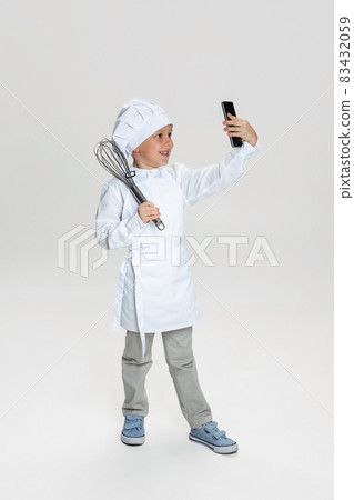 Close-up portrait of little happy boy in white cook uniform and huge chef's hat using phone isolated on white studio background. Close-up portrait of little happy boy in white cook uniform and huge chef's hat using phone isolated on white studio background. 83432059