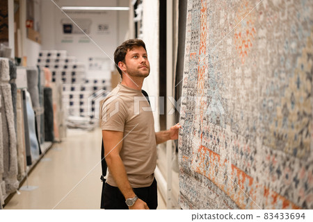 Handsome casual guy looking at rack and exploring textile of hanging carpet while shopping in modern center Handsome casual guy looking at rack and exploring textile of hanging carpet while shopping in modern center 83433694