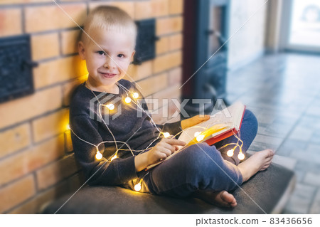 Christmas background. The boy is sitting near the fireplace with a book. Child with yellow lights. Waiting for the new year. Christmas illumination. Brick wall. Bright smile Christmas background. The boy is sitting near the fireplace with a book. Child with yellow lights. Waiting for the new year. Christmas illumination. Brick wall. Bright smile 83436656