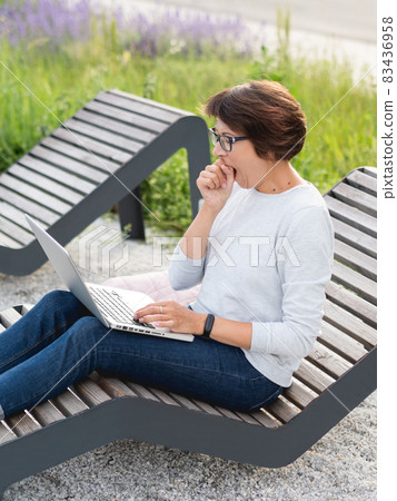 Woman sits with laptop on urban park bench. Freelancer yawns at work. Student learns remotely from outdoors. Modern lifestyle. 83436958