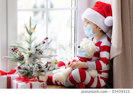 Happy child wearing Christmas pajamas sitting on windowsill Happy child wearing Christmas pajamas sitting on windowsill 83439323
