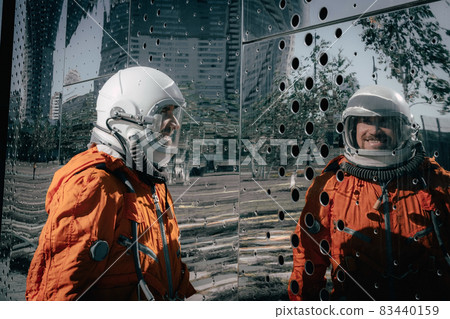 Portrait of smiling astronaut wearing orange spacesuit and space helmet near an abstract mirror wall. Portrait of smiling astronaut wearing orange spacesuit and space helmet near an abstract mirror wall. 83440159