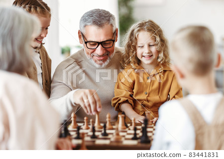 Children brother and sister playing chess while sitting in living room with senior grandparents Children brother and sister playing chess while sitting in living room with senior grandparents 83441831