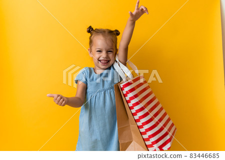 Portrait Beautiful Happy Little Preschool Girl Smiling Cheerful Holding Cardboard Bags Points Finger To Side And Up On Orange Yellow Background. Happiness, Consumerism, Sale People Shopping Concept 83446685