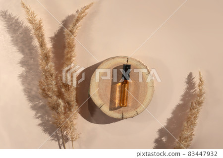 Glass perfume sample with transparent brown liquid on a wooden tray lying on a beige background with pampas grass. Luxury and natural cosmetics presentation. Tester on woodcut in sunlight. Top view 83447932
