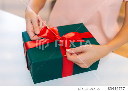 Close-up top view of unrecognizable female hands wrapping Christmas gift box tying red ribbon and decorating beautiful bow, closeup. 83456757