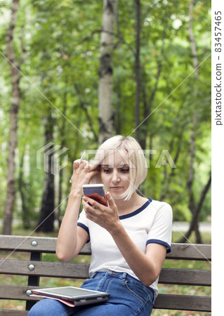 Beautiful young girl with short blond hair is watching a message in a mobile phone. She has a tablet on her lap Beautiful young girl with short blond hair is watching a message in a mobile phone. She has a tablet on her lap 83457465