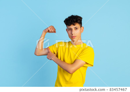 Full-length portrait of young boy in yellow T-shirt touching biceps isolated over blue background. Sport motivation Full-length portrait of young boy in yellow T-shirt touching biceps isolated over blue background. Sport motivation 83457467