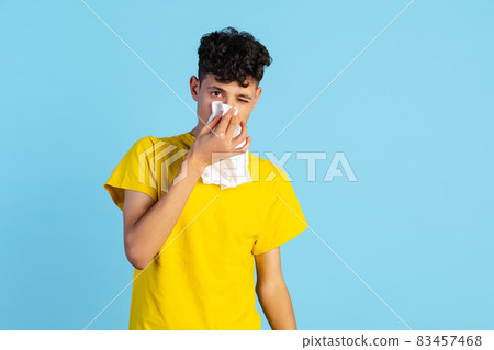 Close-up portrait of young boy in yellow T-shirt sneezing isolated over blue background. Health care Close-up portrait of young boy in yellow T-shirt sneezing isolated over blue background. Health care 83457468