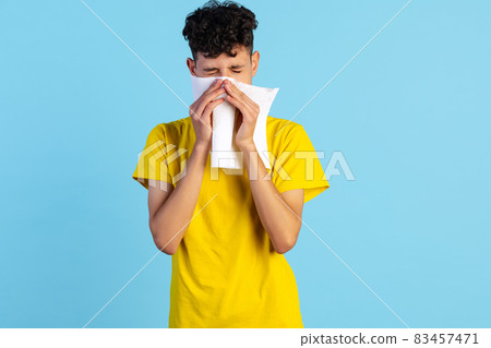 Close-up portrait of young boy in yellow T-shirt sneezing isolated over blue background. Health care Close-up portrait of young boy in yellow T-shirt sneezing isolated over blue background. Health care 83457471