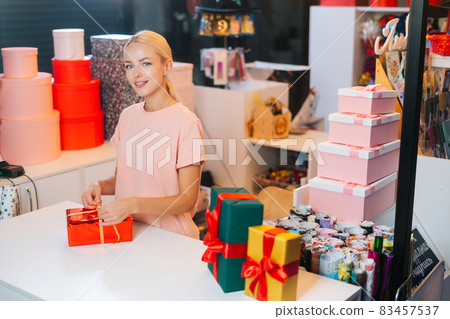 Attractive young saleswoman holding and using golden ribbon to tie bow for red wrapped gift box for Christmas present at counter of holiday shop. Attractive young saleswoman holding and using golden ribbon to tie bow for red wrapped gift box for Christmas present at counter of holiday shop. 83457537
