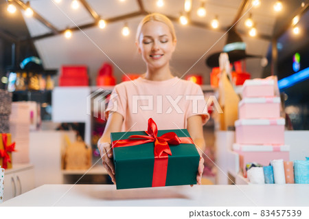 Focus on foreground of young sales woman offering take Christmas gift box tied red ribbon and decorated beautiful bow at counter of holiday store. Focus on foreground of young sales woman offering take Christmas gift box tied red ribbon and decorated beautiful bow at counter of holiday store. 83457539