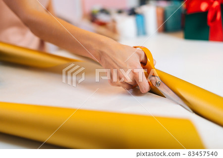 Close up of unrecognizable young woman cutting with scissors wrapping golden paper to package festive cardboard box with Christmas gifts. 83457540