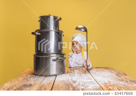 Close-up cute little boy in white cook uniform and huge chef's hat at kids kitchen with big pans isolated on yellow studio background. Close-up cute little boy in white cook uniform and huge chef's hat at kids kitchen with big pans isolated on yellow studio background. 83457608