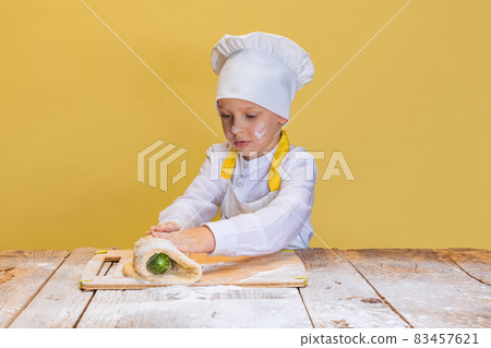 One cute boy wearing in white cook, chef jacket and hat kneads dough isolated on yellow studio background. One cute boy wearing in white cook, chef jacket and hat kneads dough isolated on yellow studio background. 83457621